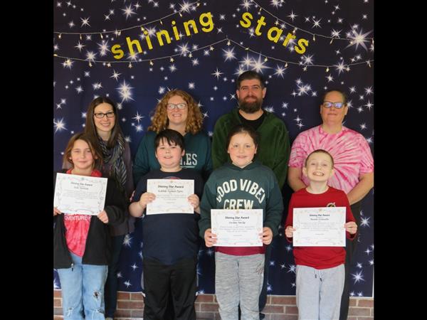 Children proudly display certificates in front of a wall.