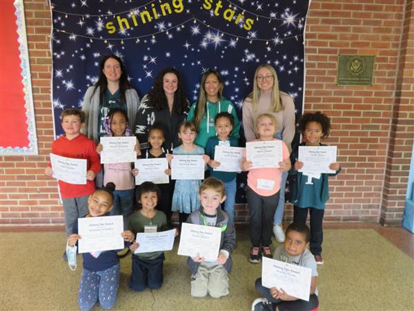 Children proudly display certificates in front of a wall.