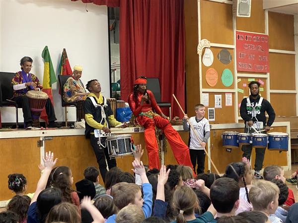 Children sitting in classroom listening to man on stage.