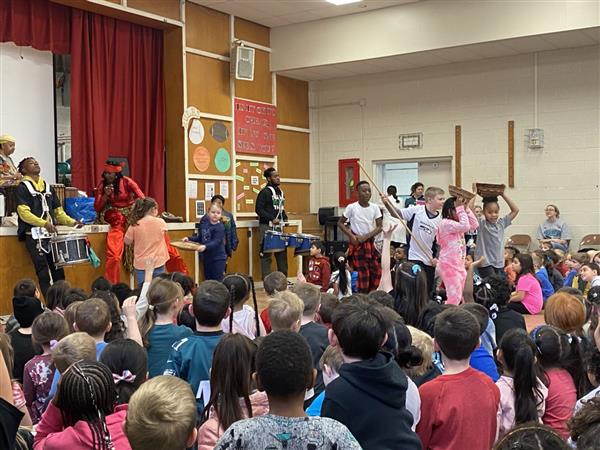 Children sitting in classroom listening to man on stage.