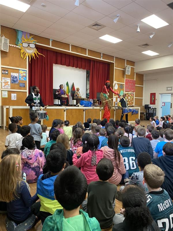 Children sitting in classroom listening to man on stage.