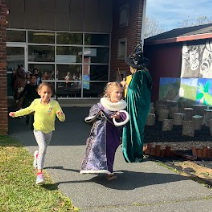 Two kids running in front of a building with a man in witch costume.