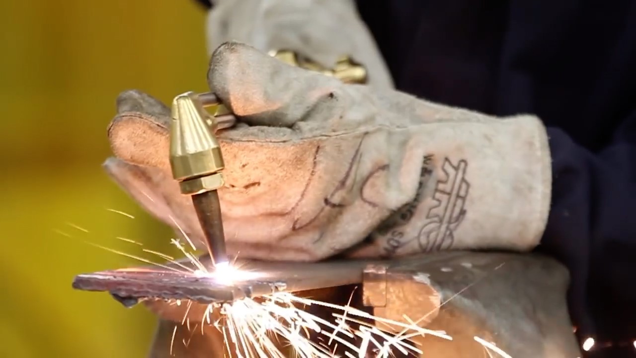 A person skillfully welding with a torch inside a factory, surrounded by industrial equipment and safety gear.