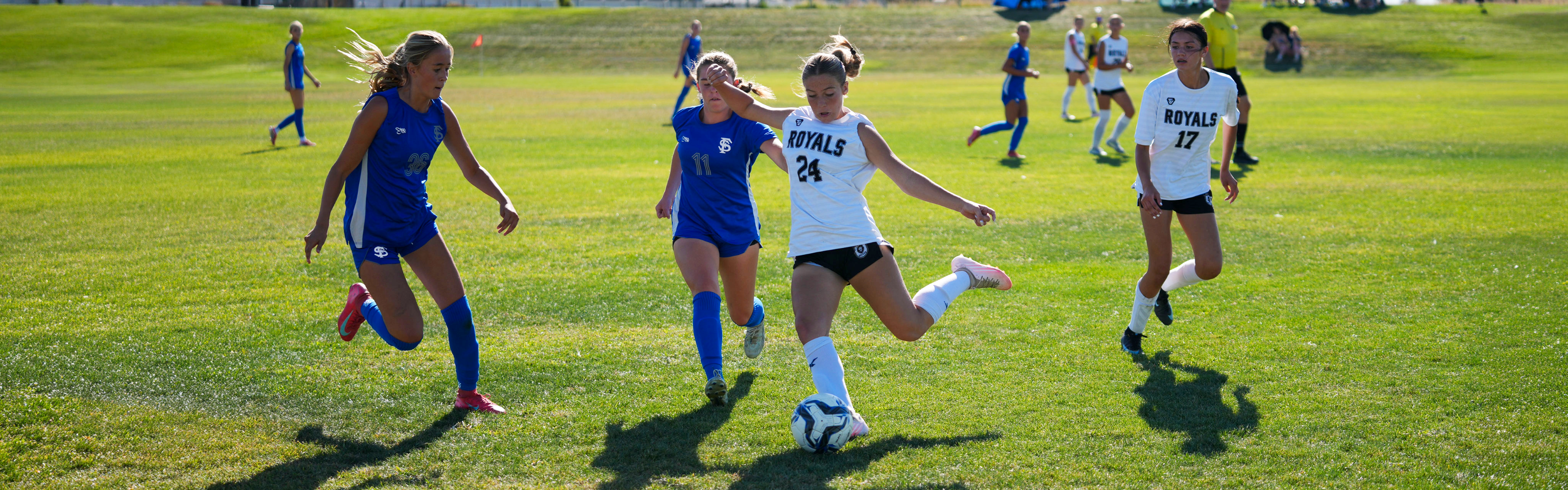 A Roy High School soccer player in a white uniform kicks the soccer ball downfield during an outdoor match as opposing defenders press in.