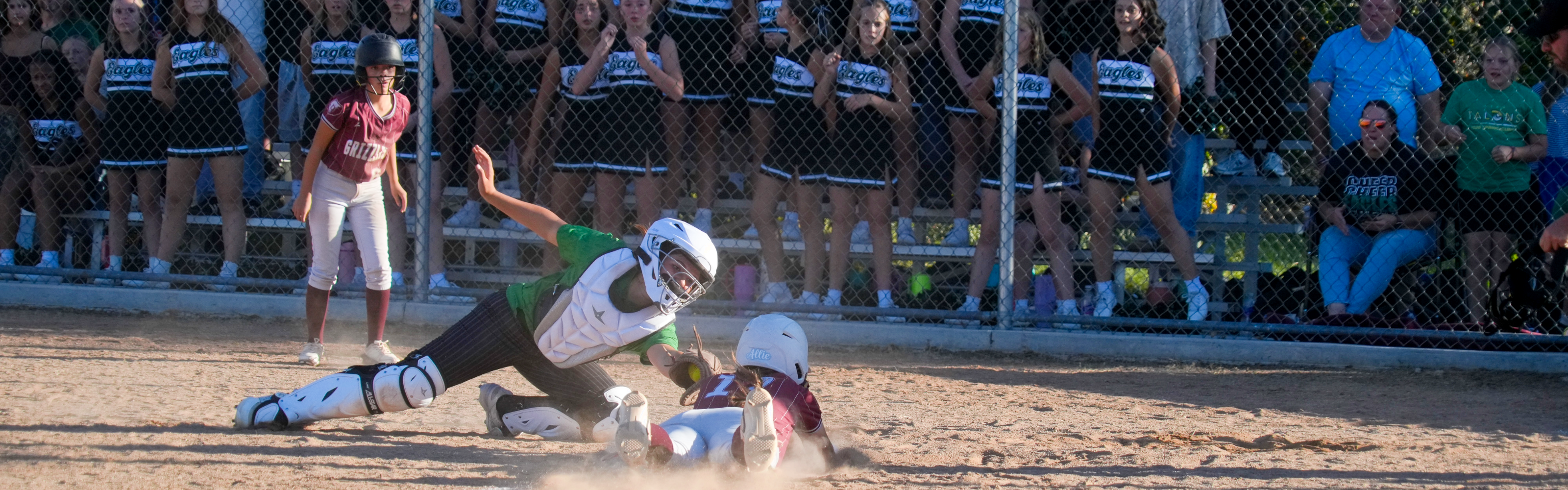 Image 4: Wide banner image highlighting an intense play at home plate during a junior high softball game as a runner slides into home while the catcher attempts a tag.   