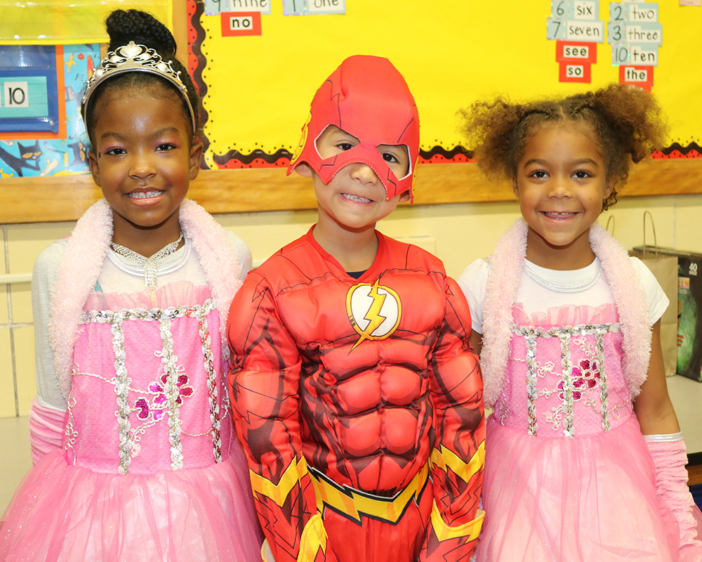Three children in costumes, posing for a photo in front of a classroom.