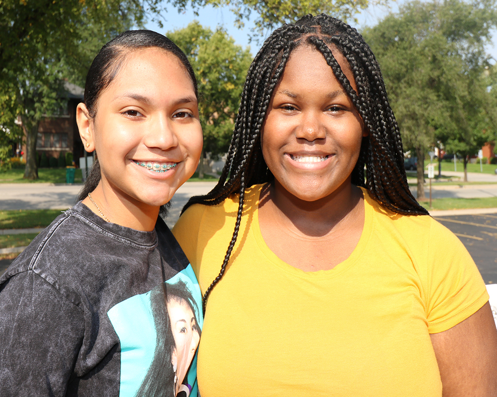 A young woman in a yellow shirt and black hairstyle stands next to a smiling girl in a white tank top, both with a backdrop of trees.