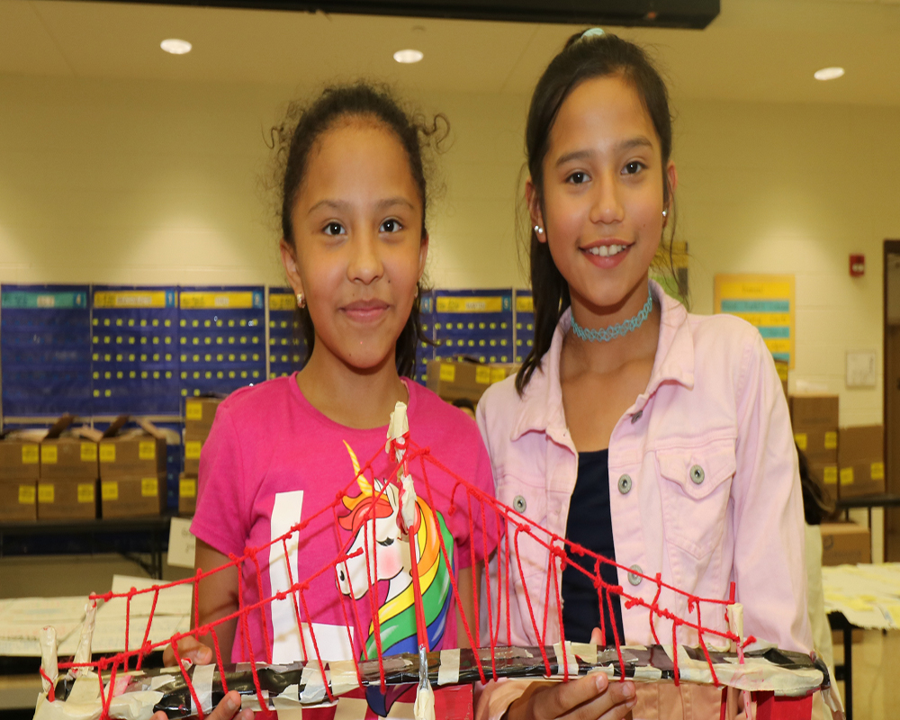 Two children standing beside a bridge made from popsicle sticks.