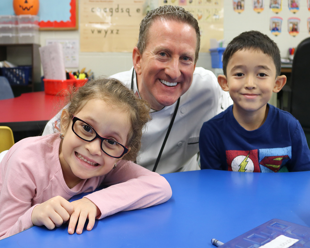 A person is standing at a table with two children, all smiling for the camera. The individual appears to be a teacher or caregiver based on their attire.