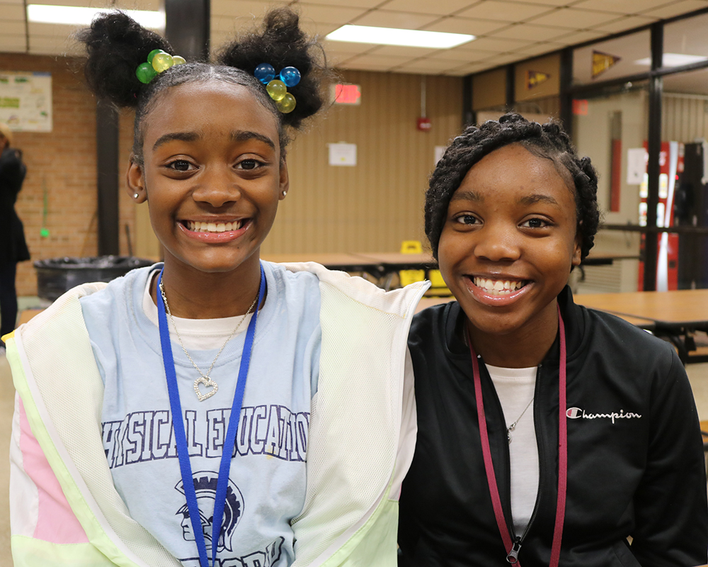Two young girls, possibly friends or sisters, posing for a photo with big smiles against a school backdrop.
