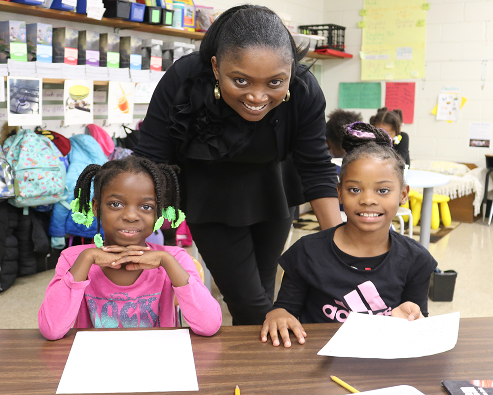 A teacher and two students at a desk in a classroom.