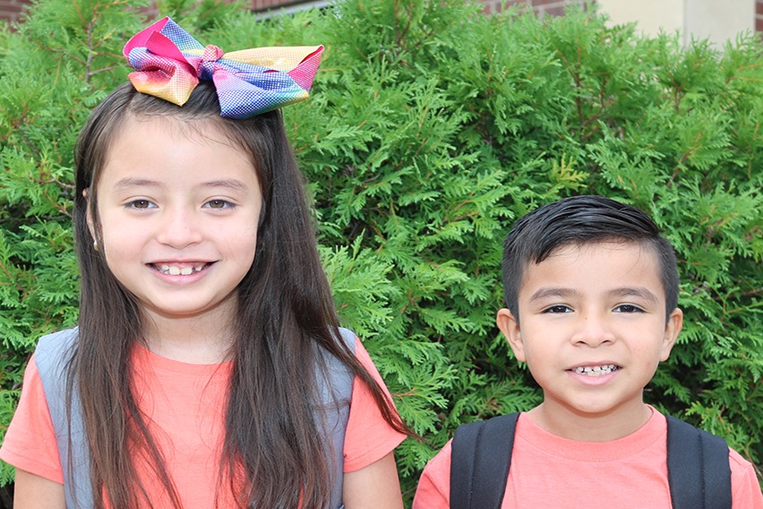 Two children, a girl and a boy, posing for a photo in front of a green shrub.