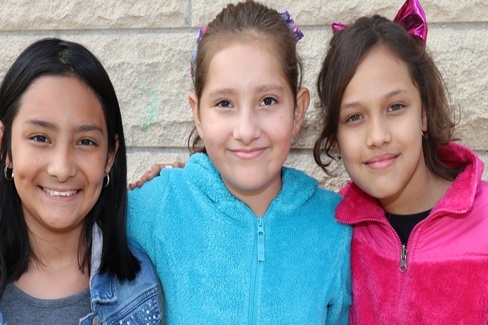 Three young girls posing together for a photo, each wearing different color jackets and smiles.