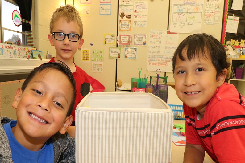 Two children and a man standing in front of a colorful educational toy at a desk in a classroom.