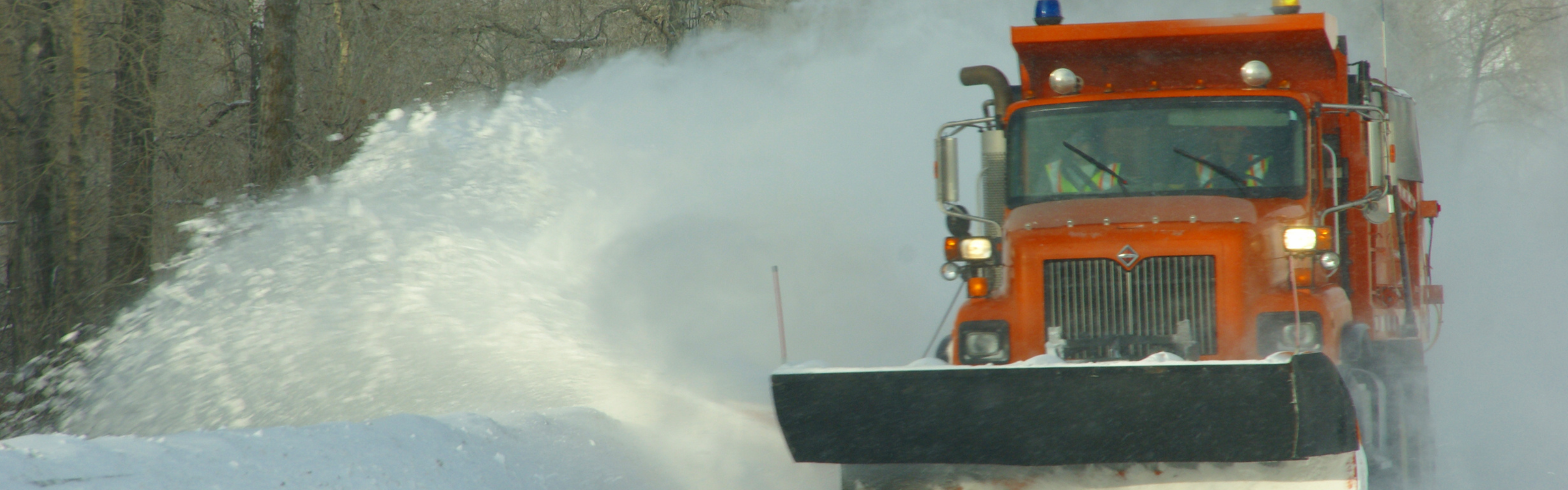 photo of a orange snow plow plowing snow on a road 
