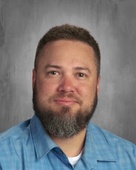 Man with dark short hair and dark beard wearing a blue t-shirt with a grey background