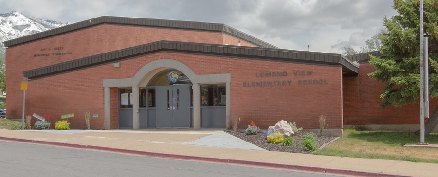 Exterior front entrance of Lomond View Elementary School, featuring a single-story red brick building with the school name on the facade, a blue mascot logo above the arched entryway, small landscaped garden beds along the sidewalk, and snow-dusted mountains rising in the background.