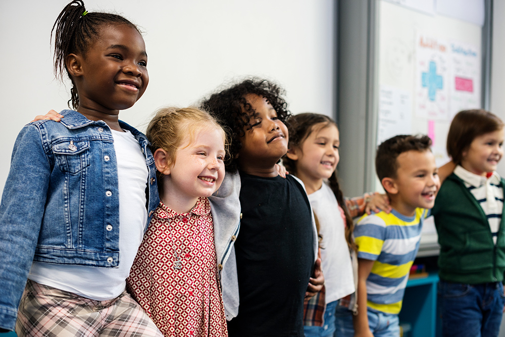 A group of six diverse young children stand together in a row, smiling and posing with their arms around one another in a brightly lit school setting. The children are dressed in casual clothes, including a denim jacket, a red patterned dress, and striped shirts. They appear happy and are looking off-camera towards the left.