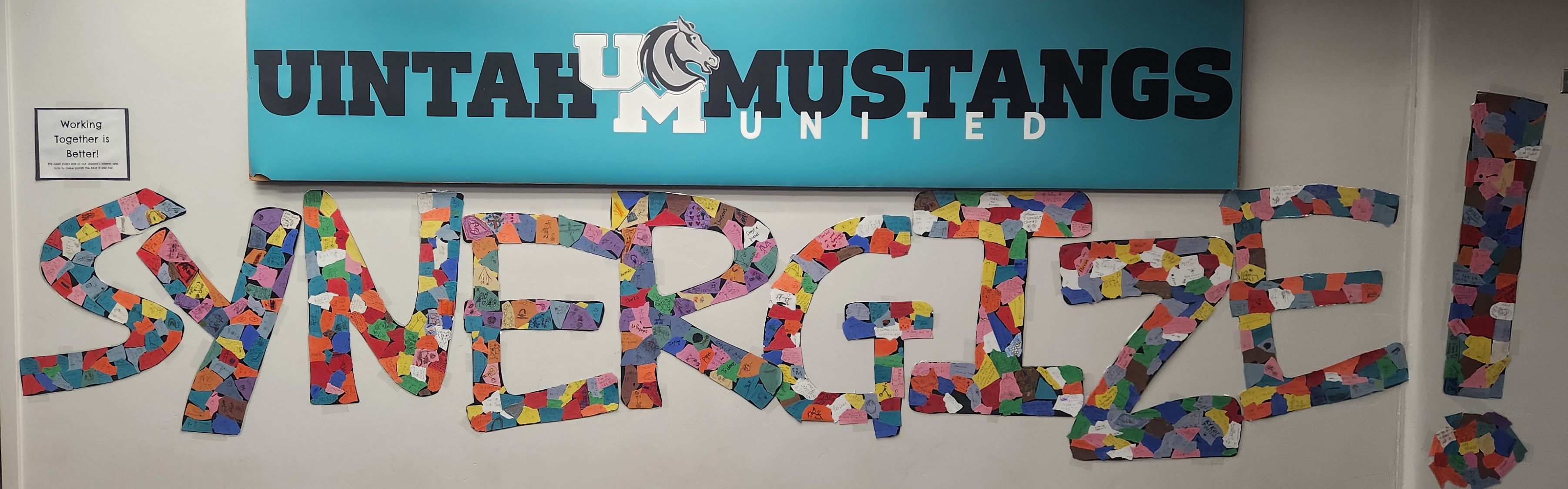 Picture of the school hallway.  A blue sign reads 'Uintah Mustangs, United" with the Uintah logo  of the letters U and M with a mustang head.  Below is a mural of the word 'synergize' created by students. 