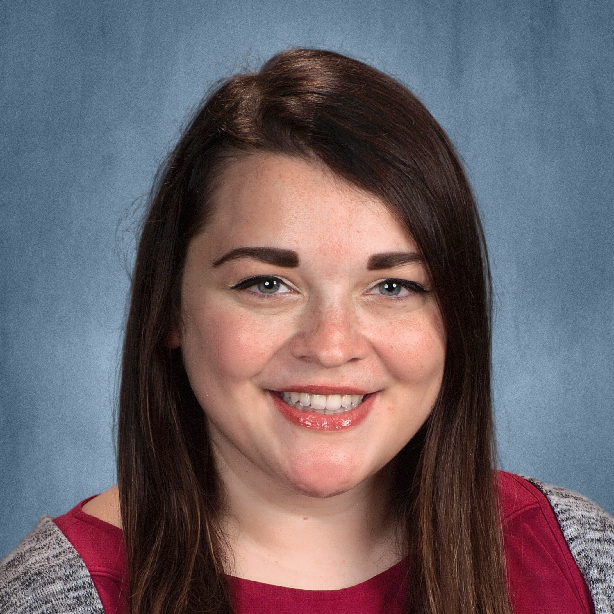 Professional headshot of head choir director Bridgette Pineau, smiling with long brown hair against a neutral blue background