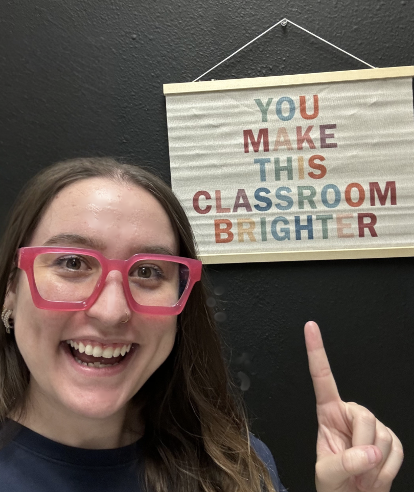 Teacher standing beside and pointing to a theatre program sign in a classroom.