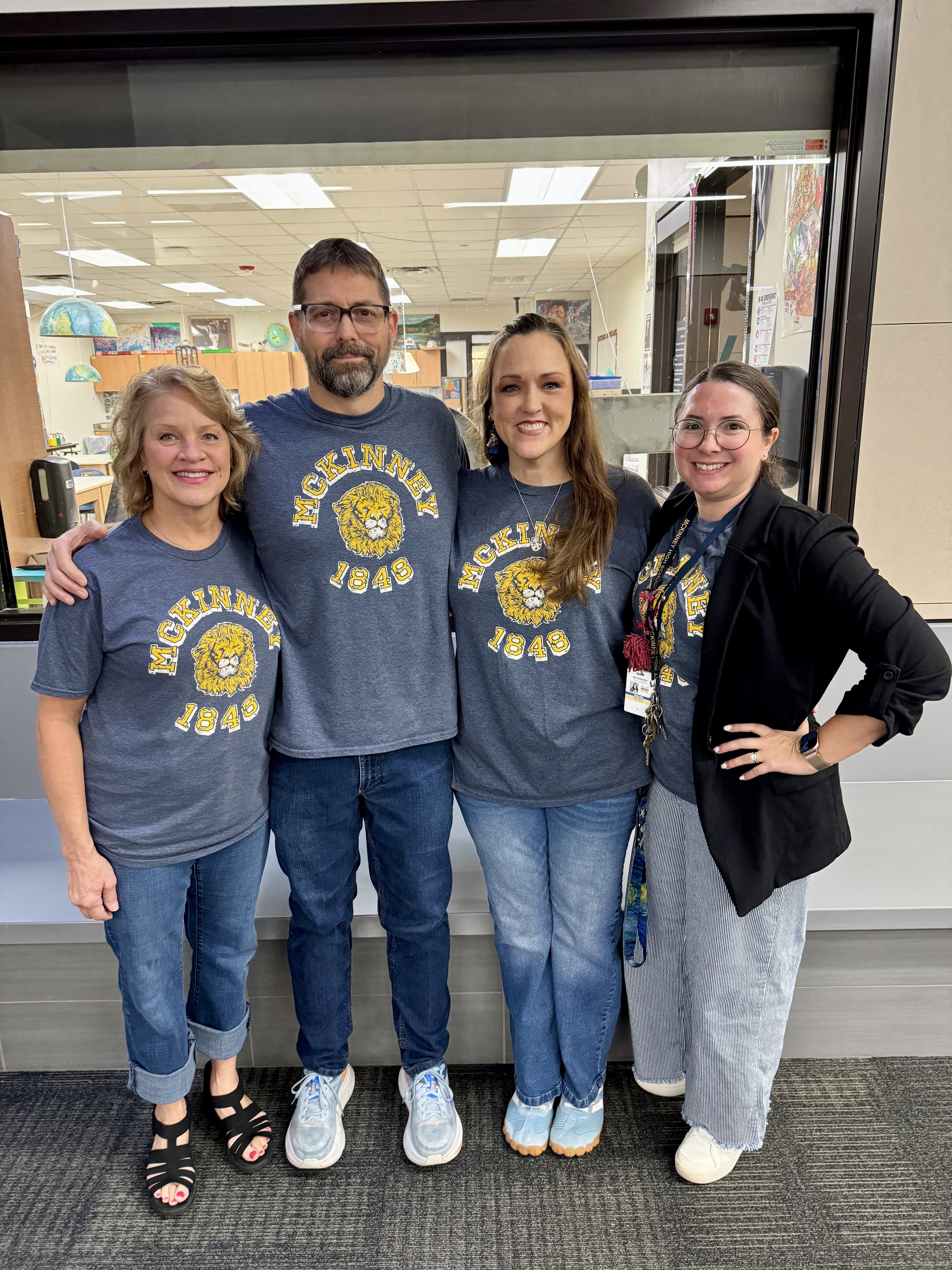A group of McKinney High School art teachers wearing matching blue t-shirts, smiling and posing together in a school hallway.