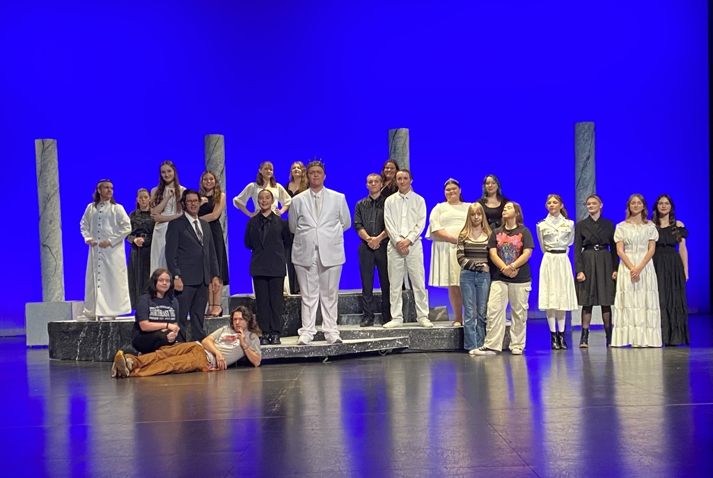 A High School Theatre group posing in their costumes for the play Antigone against a blue-lit stage