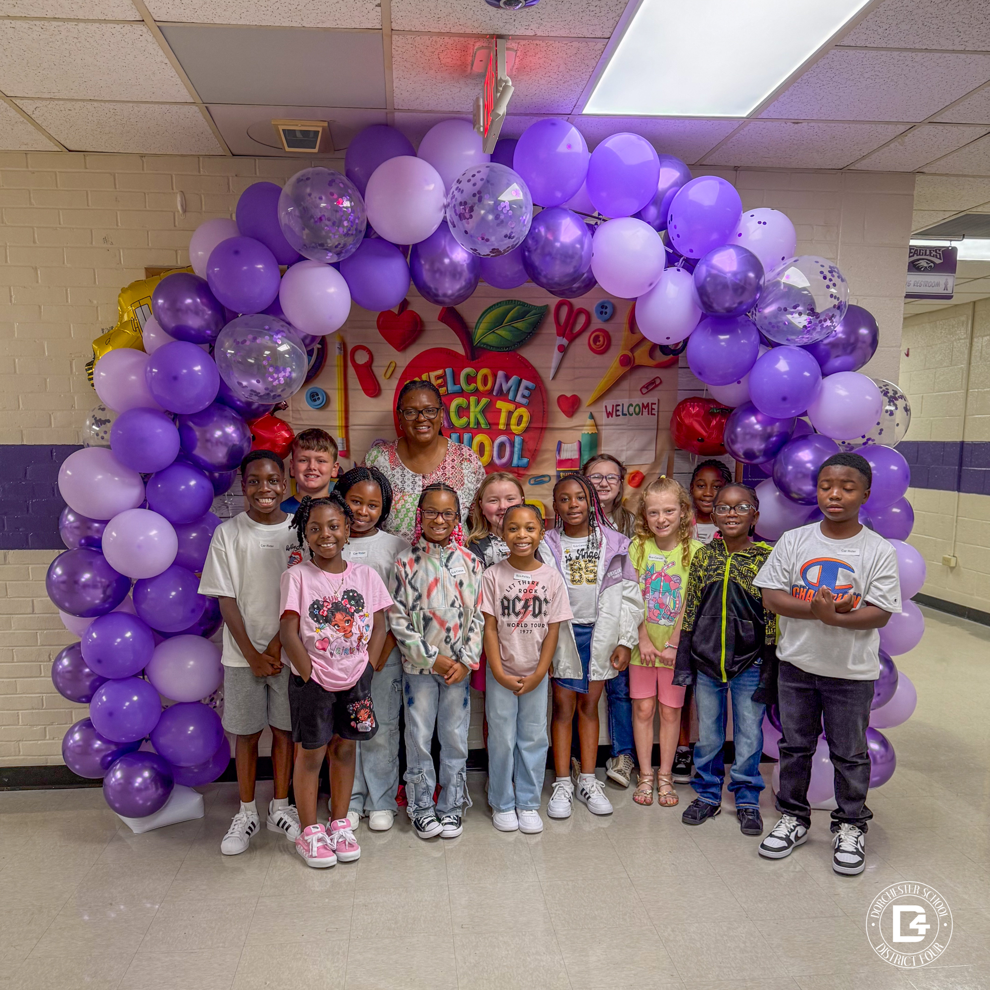  A group of elementary students from Clay Hill Elementary pose with a teacher under a festive purple balloon arch. Behind them is a colorful "Welcome Back to School" banner featuring school-themed decorations like scissors, pencils, and glue. The group is smiling and standing in a hallway with purple and white walls. The Dorchester School District Four logo is in the corner of the image.