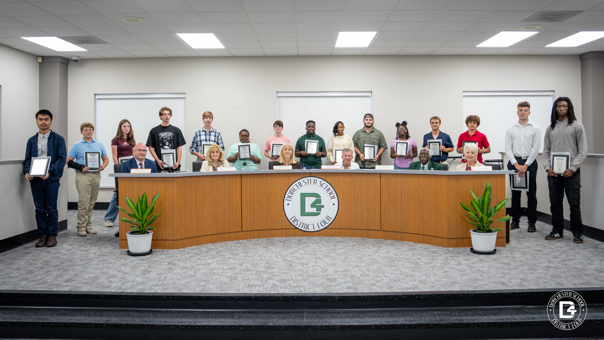 A large group photo inside the Dorchester School District Four boardroom. Students stand in a row behind the board table holding framed certificates, while district leaders sit at the front of the table. The district logo is displayed on the wooden panel at the center of the table, and two potted plants are placed on each side.