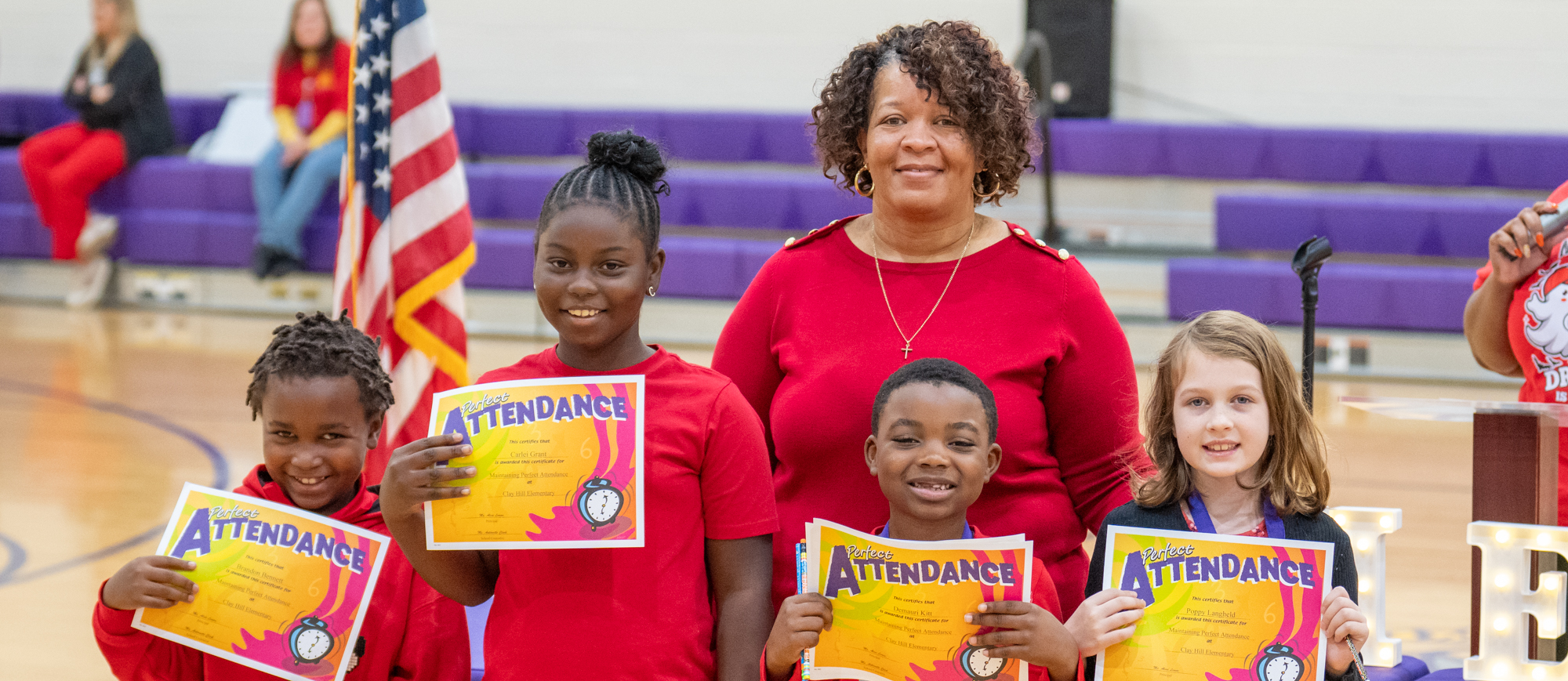 Four smiling students stand in front of a teacher during an award ceremony in a gym. Each student holds a bright orange and yellow “Perfect Attendance” certificate and wears a medal. The teacher stands proudly behind them, and an American flag and “CHES” marquee letters are visible in the background.