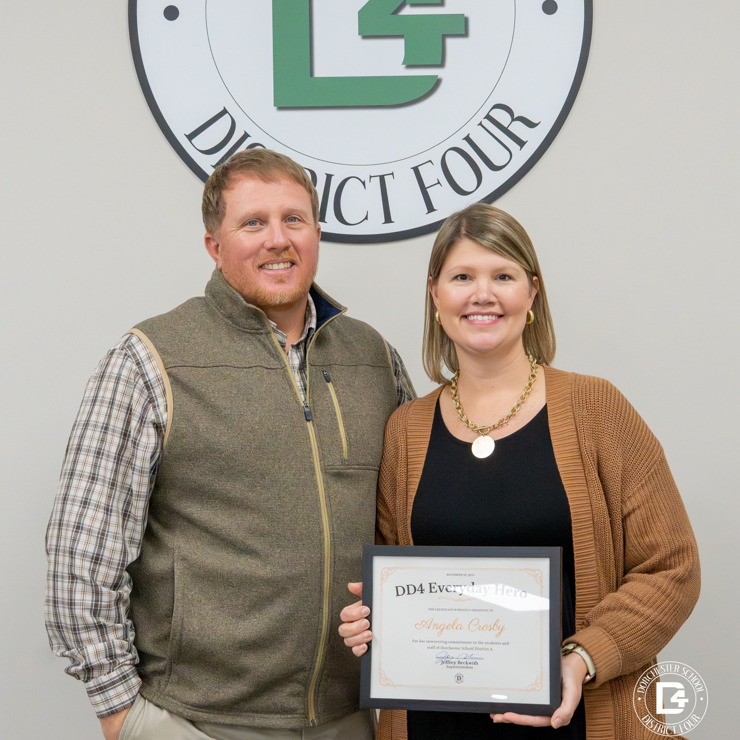 Angela Crosby stands beside her husband in a posed photo inside a Dorchester School District Four office, smiling toward the camera beneath the district seal. Angela holds a DD4 Everyday Hero certificate, and the image reflects pride, support, and shared celebration of her recognition.