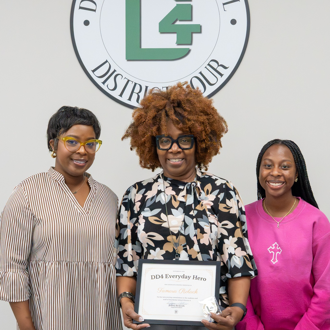 Tamara Rolack stands centered in a posed photo inside a Dorchester School District Four office, holding a DD4 Everyday Hero certificate. She is flanked by two family members, all smiling toward the camera beneath the district seal on the wall. The image reflects pride, support, and shared celebration of her recognition.
