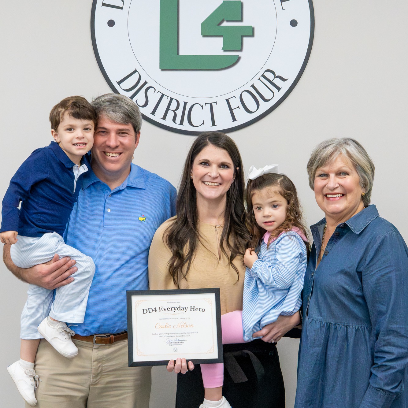 Carlie Nelson stands with her family inside a Dorchester School District Four office, smiling beneath the district seal. She holds a DD4 Everyday Hero certificate while family members stand close, including children in her arms. The photo reflects pride, support, and a shared moment of celebration.