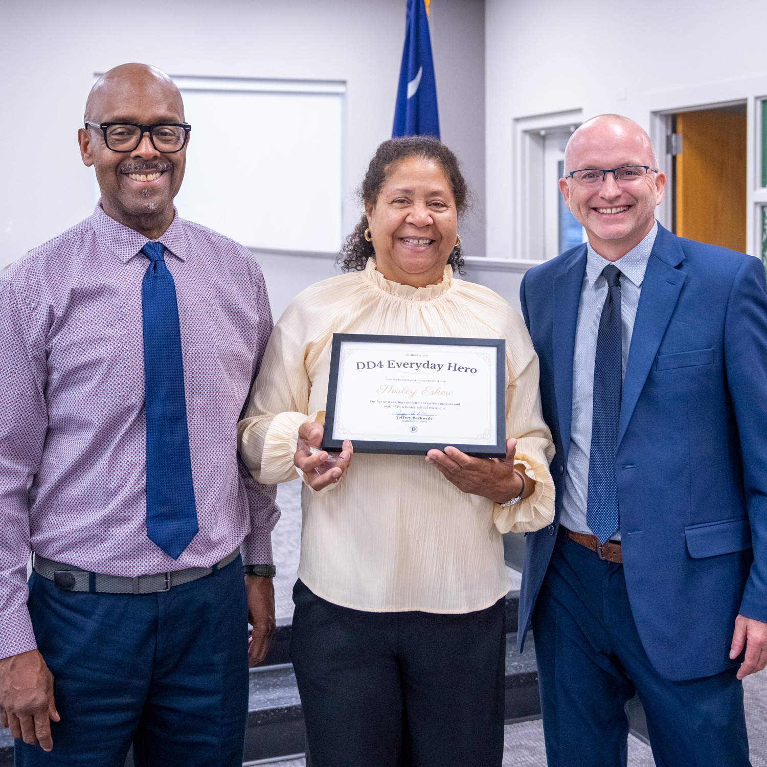 Mrs. Shirly Eskew stands centered in a district meeting room, smiling as she holds her DD4 Everyday Hero certificate. She is flanked by two district leaders, all dressed professionally, with the Dorchester School District Four setting visible behind them. The photo captures a moment of pride, recognition, and appreciation for her service.