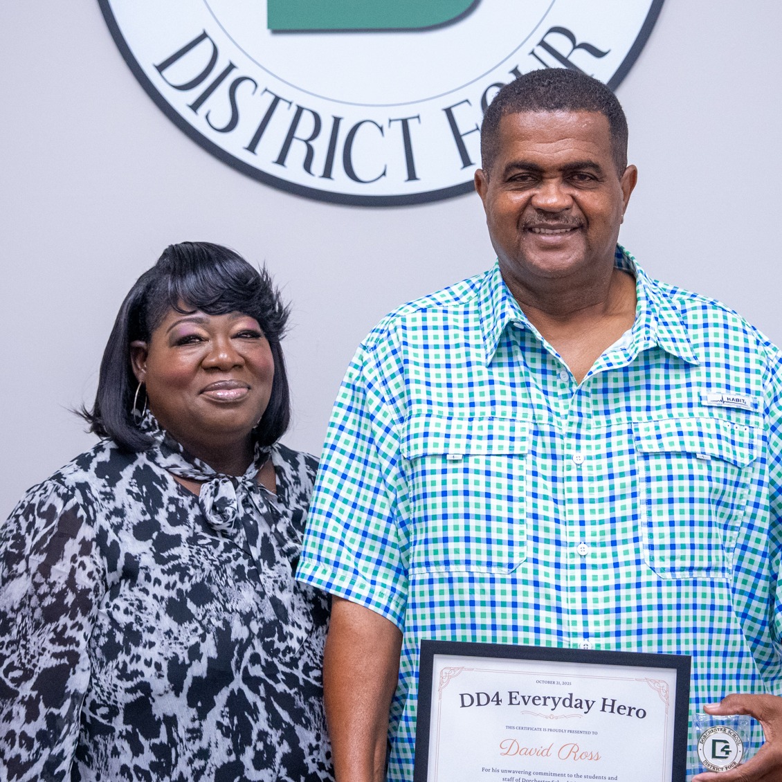 David Ross stands beside his wife in a posed photo inside a Dorchester School District Four office, smiling beneath the district seal. David holds a DD4 Everyday Hero certificate and coin, while his wife stands proudly at his side, reflecting shared pride and support for his recognition.