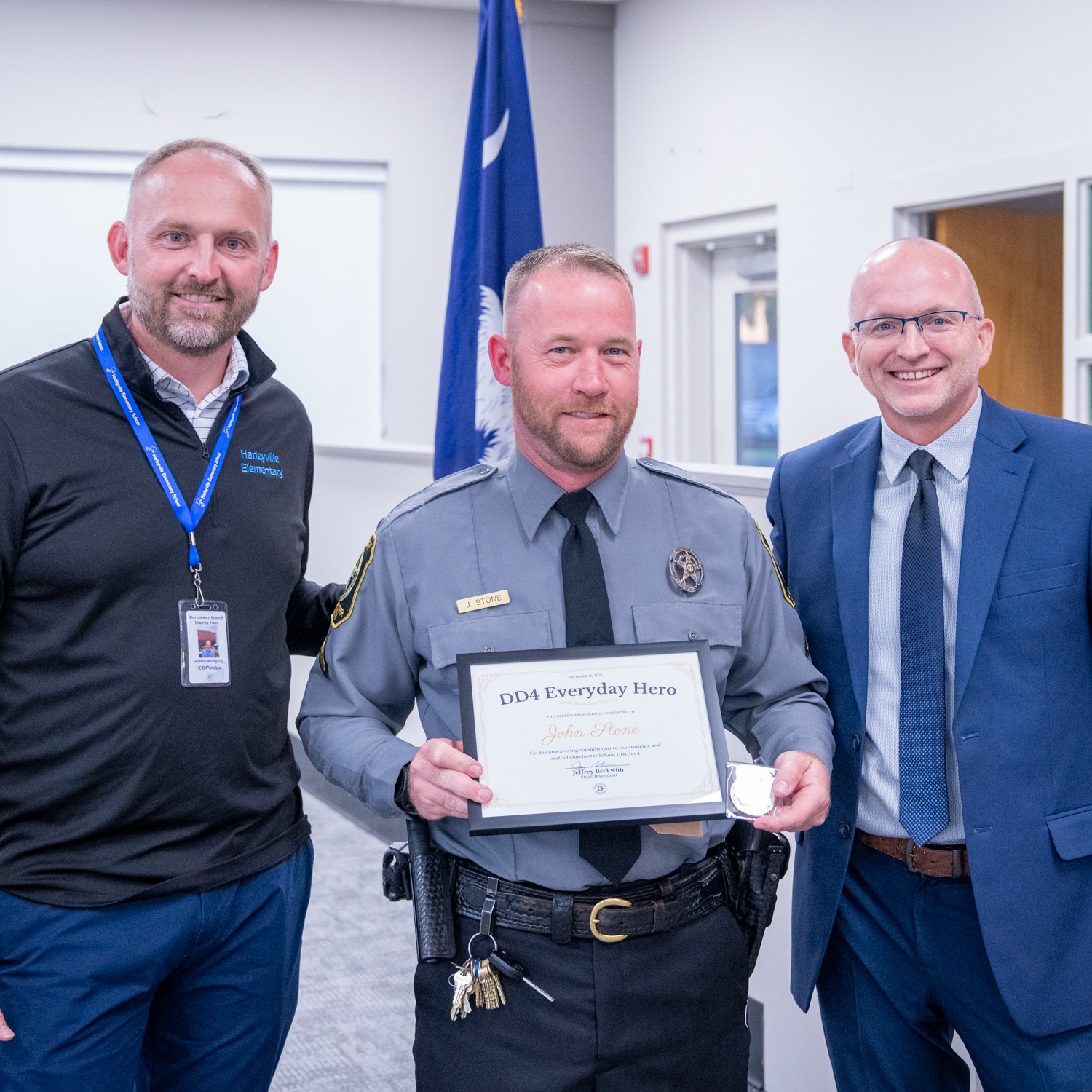 John Stone stands centered in a district meeting room, wearing his law enforcement uniform and smiling as he holds his DD4 Everyday Hero certificate and coin. He is flanked by two district leaders, all standing together in a professional setting that highlights recognition, respect, and appreciation for his service.