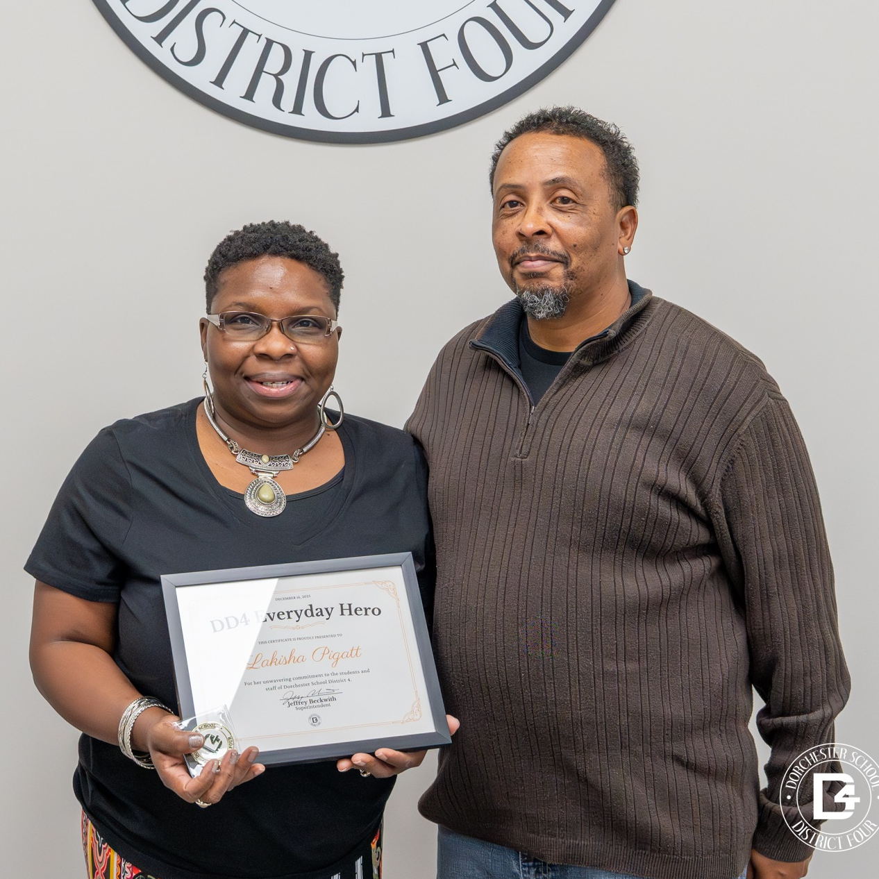 Lakisha Pigatt stands proudly beside her husband, holding her Everyday Hero certificate and coin, following her recognition by Dorchester School District Four. The pair smile warmly in front of the district seal, marking a meaningful moment of appreciation and shared pride.