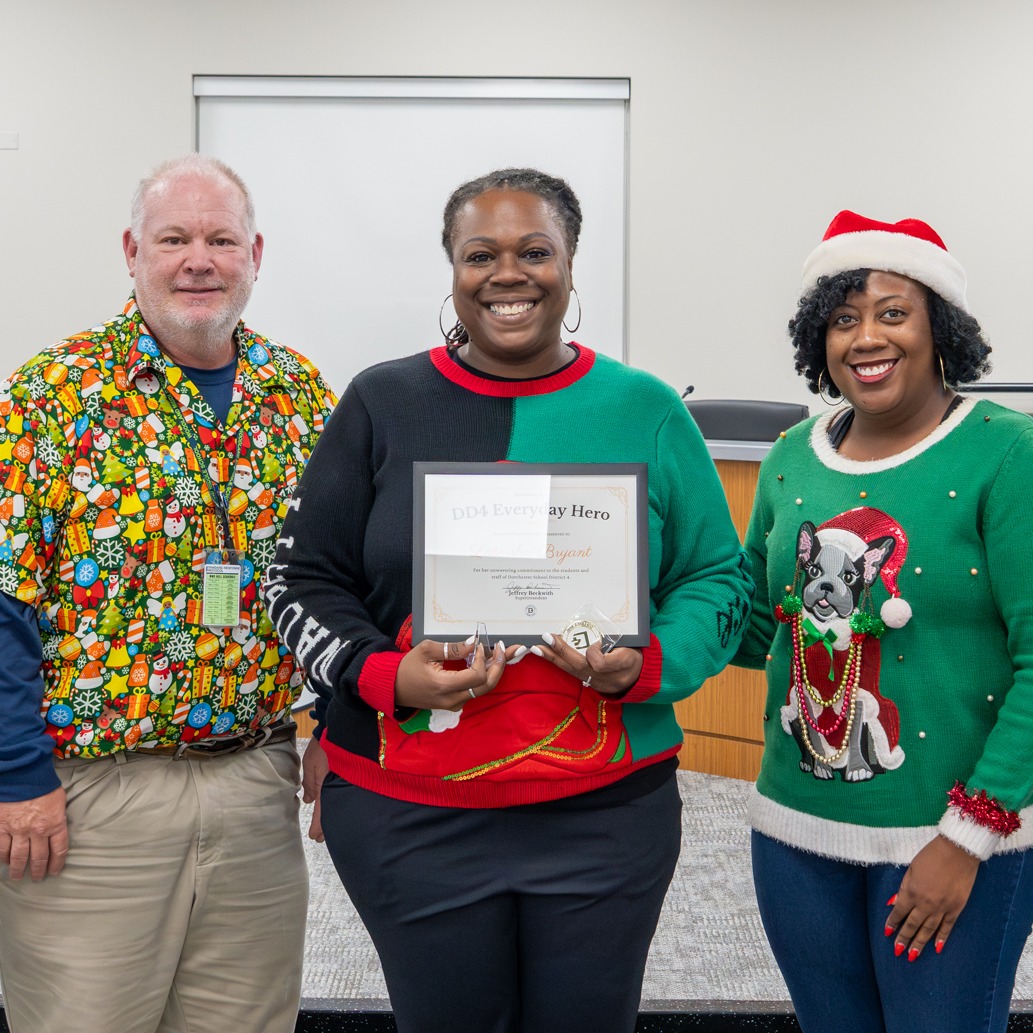 Latorsha Bryant smiles while holding her Everyday Hero certificate and coin, surrounded by district leaders and colleagues during her recognition by Dorchester School District Four. The group stands together in the boardroom, celebrating her service in a warm, festive moment.