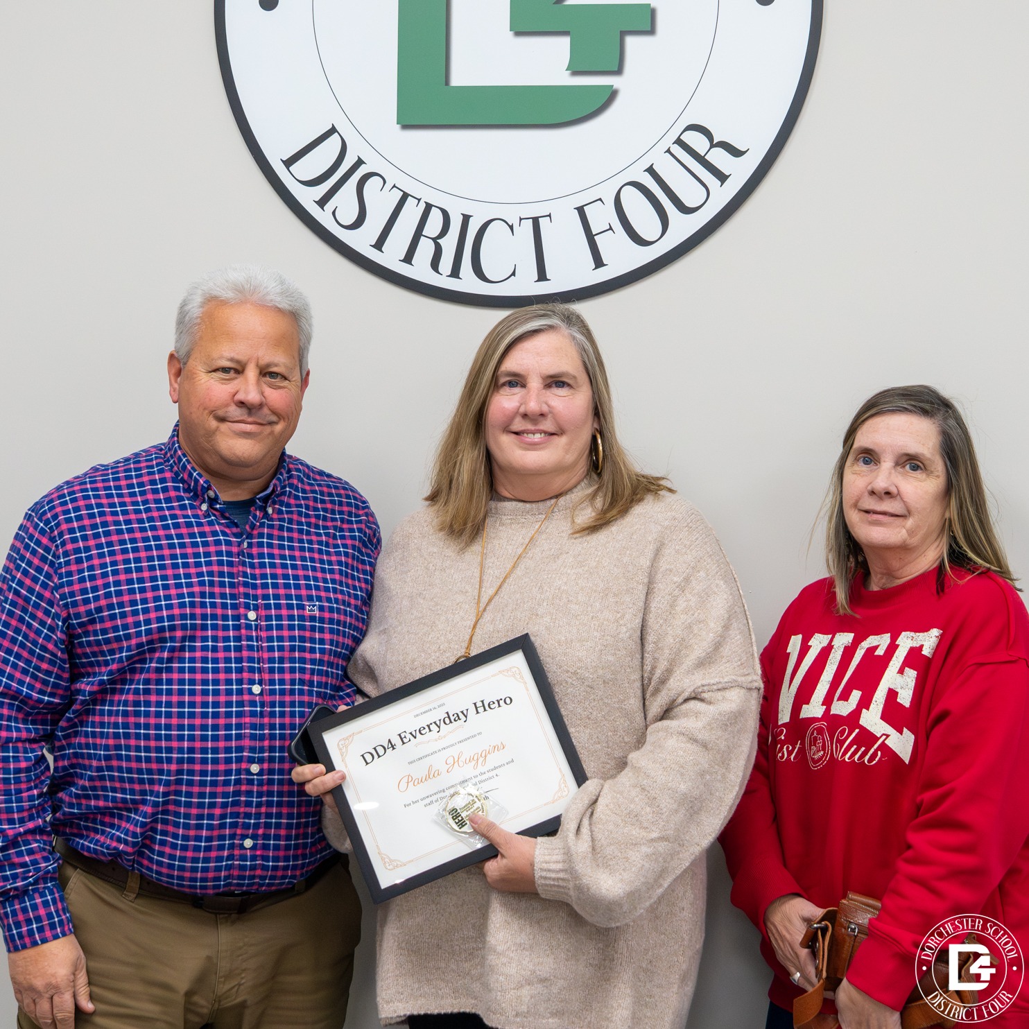 Paula Huggins stands with her family in front of the Dorchester School District Four seal, holding her Everyday Hero certificate and coin, smiling after being recognized for her service and dedication.