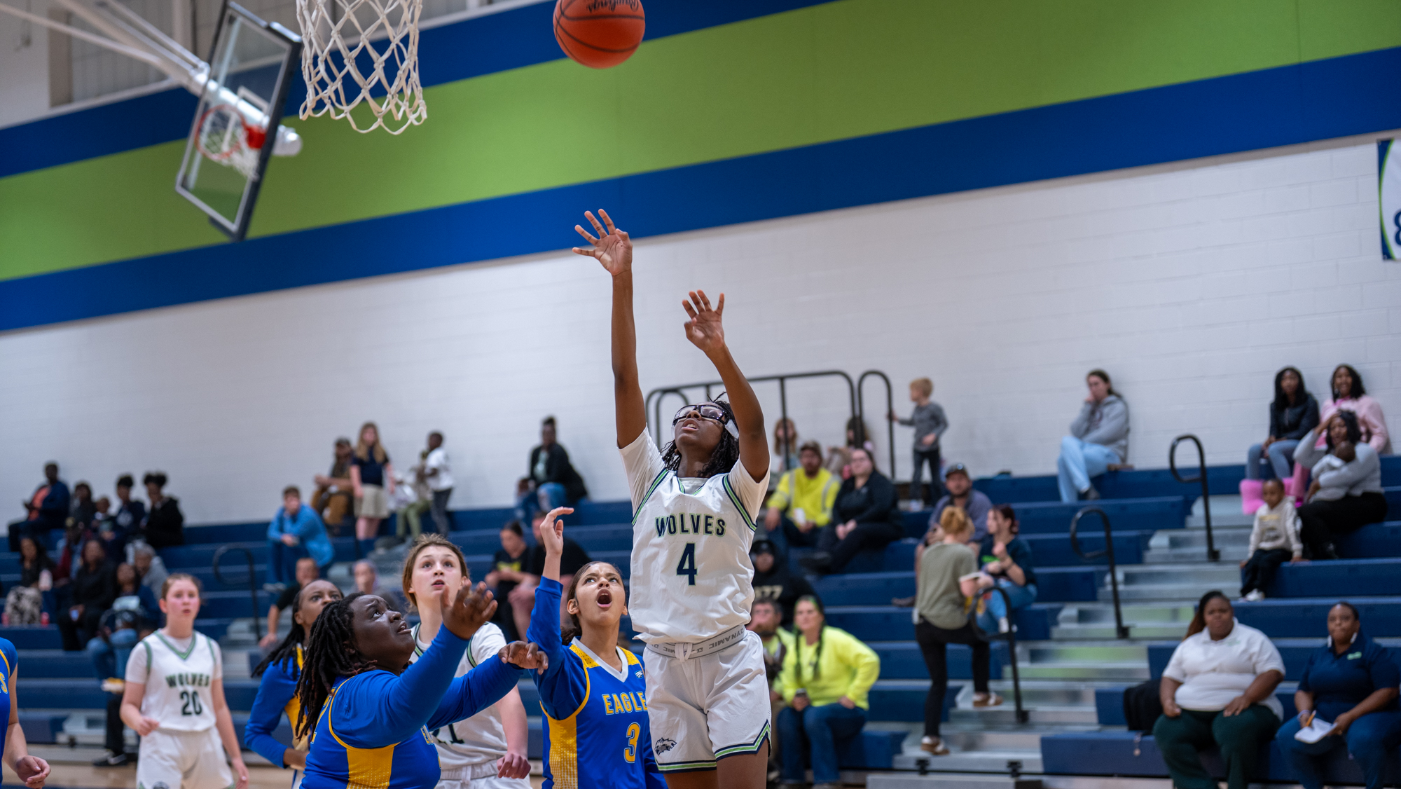Woodland Middle School girls basketball player in a white Wolves uniform (#4) jumps for a layup as the ball approaches the hoop, surrounded by defenders in blue and yellow Eagles uniforms, with teammates and spectators watching from the bleachers in the gym.