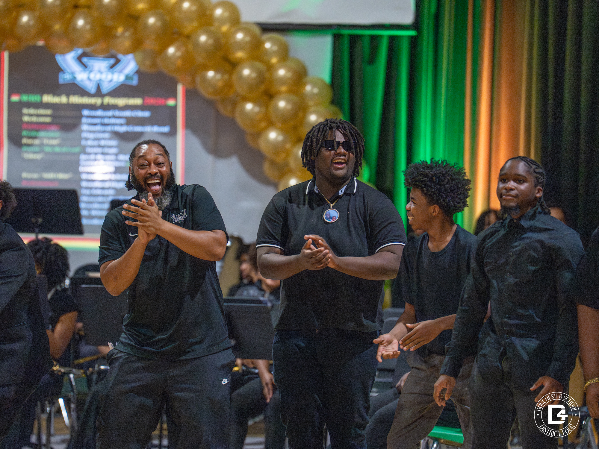 A male teacher on the far left smiles and claps while standing beside three male students dressed in black during a school Black History Month program. Gold balloons arch above them, with green and gold lighting in the background. Other students with musical instruments are seated behind them, and a Dorchester School District Four logo watermark appears in the bottom right corner.