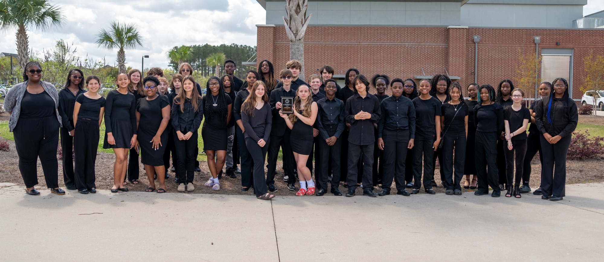 Woodland Middle School concert band members and director Ms. Yolanda Bennett pose together outside in front of the school building and palm trees. The group is dressed in black performance attire, with one student holding the award plaque at the center.