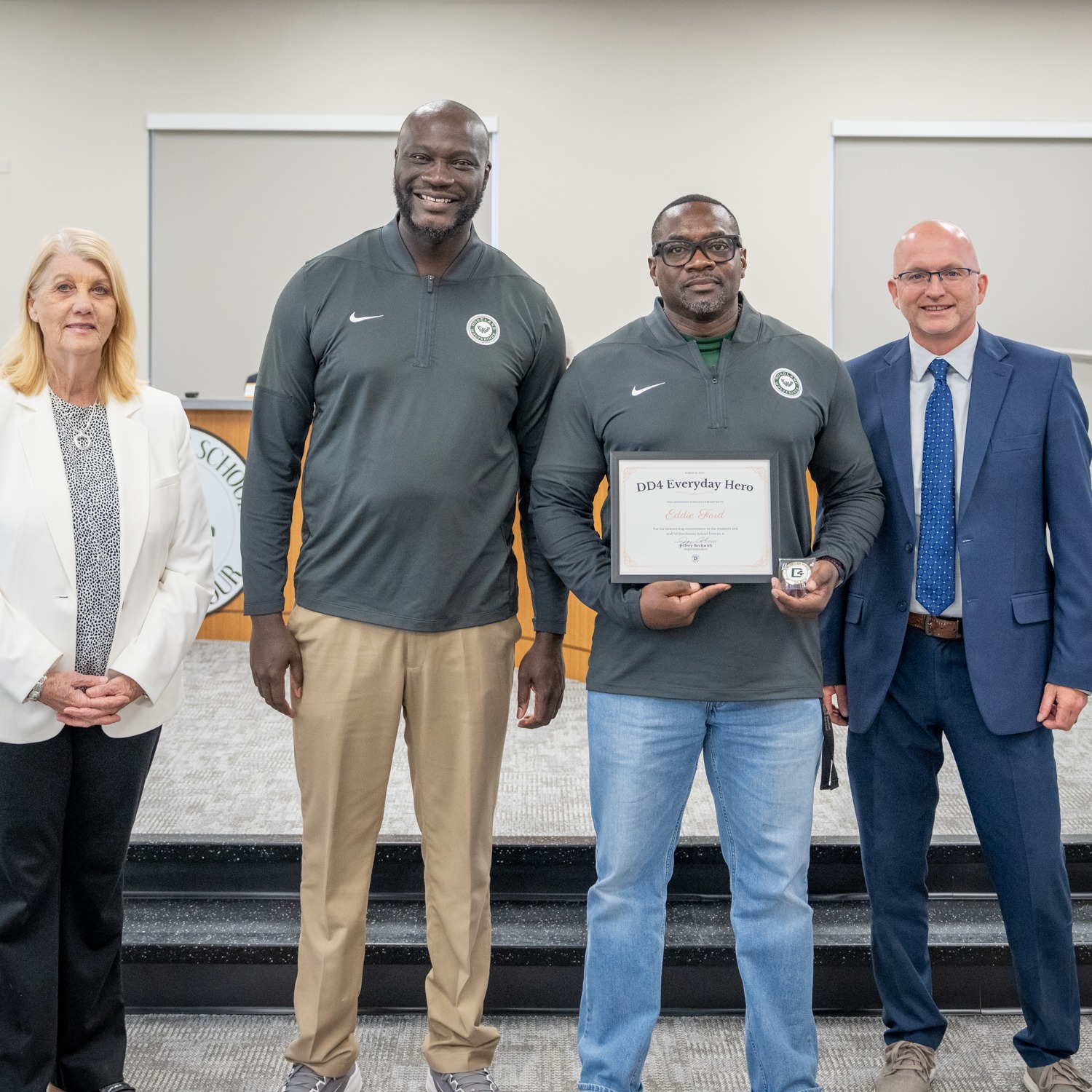 A Dorchester School District Four employee holds a framed "DD4 Everyday Hero" award and a small trophy while posing with three colleagues in the district's board room. The group stands in front of the district seal, with the South Carolina state flag visible in the background. The Dorchester School District Four logo appears in the bottom right corner.