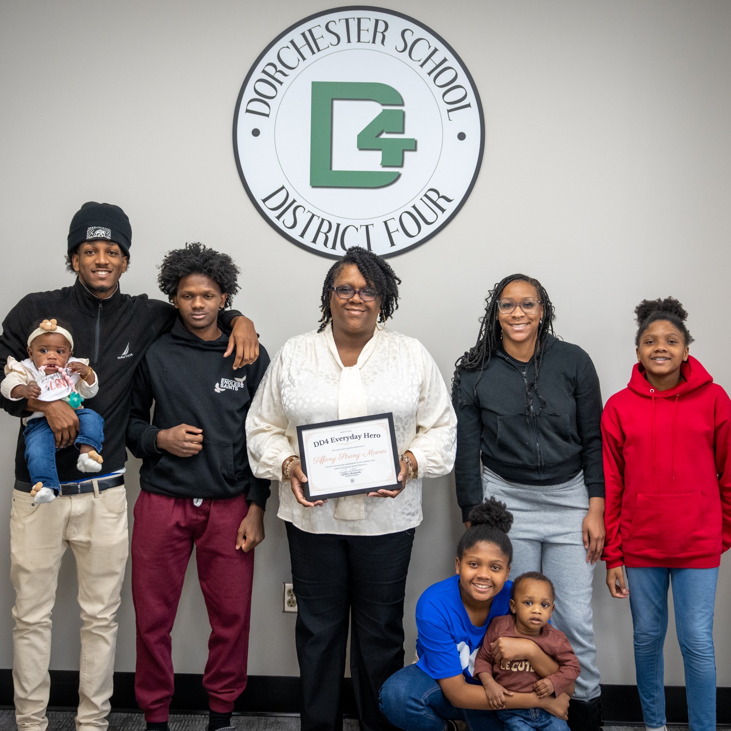 A Dorchester School District Four employee stands in front of the district's wall logo holding a framed "DD4 Everyday Hero" award, surrounded by family members including adults, children, and an infant. The group smiles together in celebration of the recognition. The Dorchester School District Four logo appears in the bottom right corner.