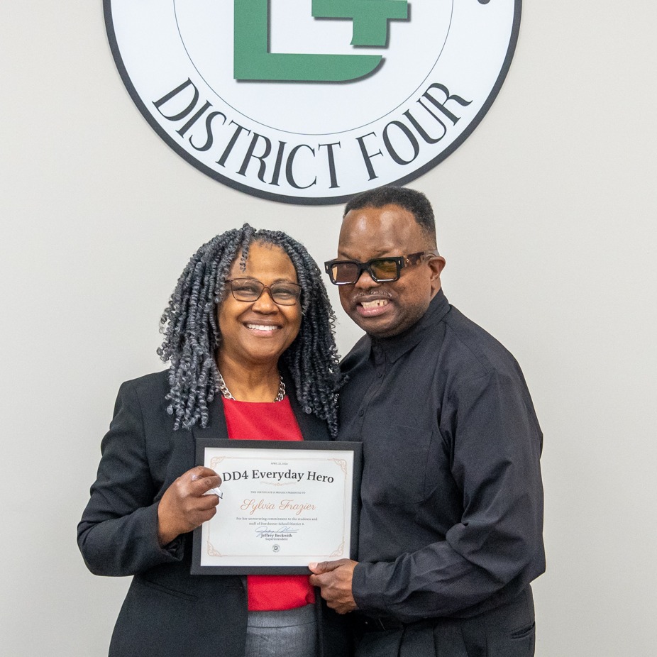 Sylvia Frazier and her Husband posing with an award under a DD4 logo on the wall behind them. 