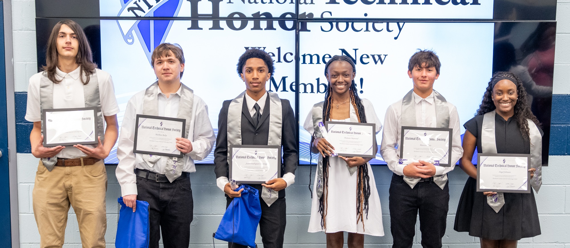 Six Woodland High School students pose proudly in front of a large National Technical Honor Society 'Welcome New Members' display screen following their induction ceremony. Each student holds a framed National Technical Honor Society certificate and a blue drawstring gift bag. The students are dressed in formal attire and wear silver NTHS honor stoles.