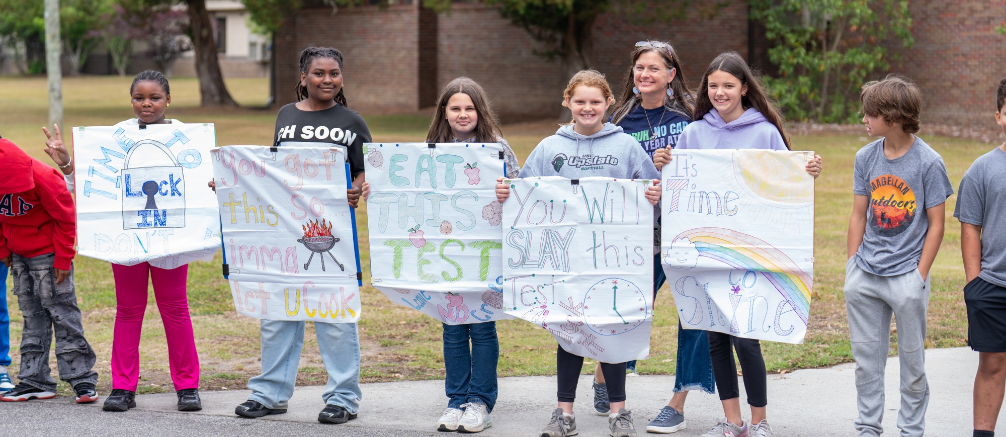 Williams Memorial Elementary students stand outside holding handmade signs with testing encouragement messages, including “Lock in,” “You got this,” “Eat this test,” “You will slay this test,” and “It’s time to shine.”