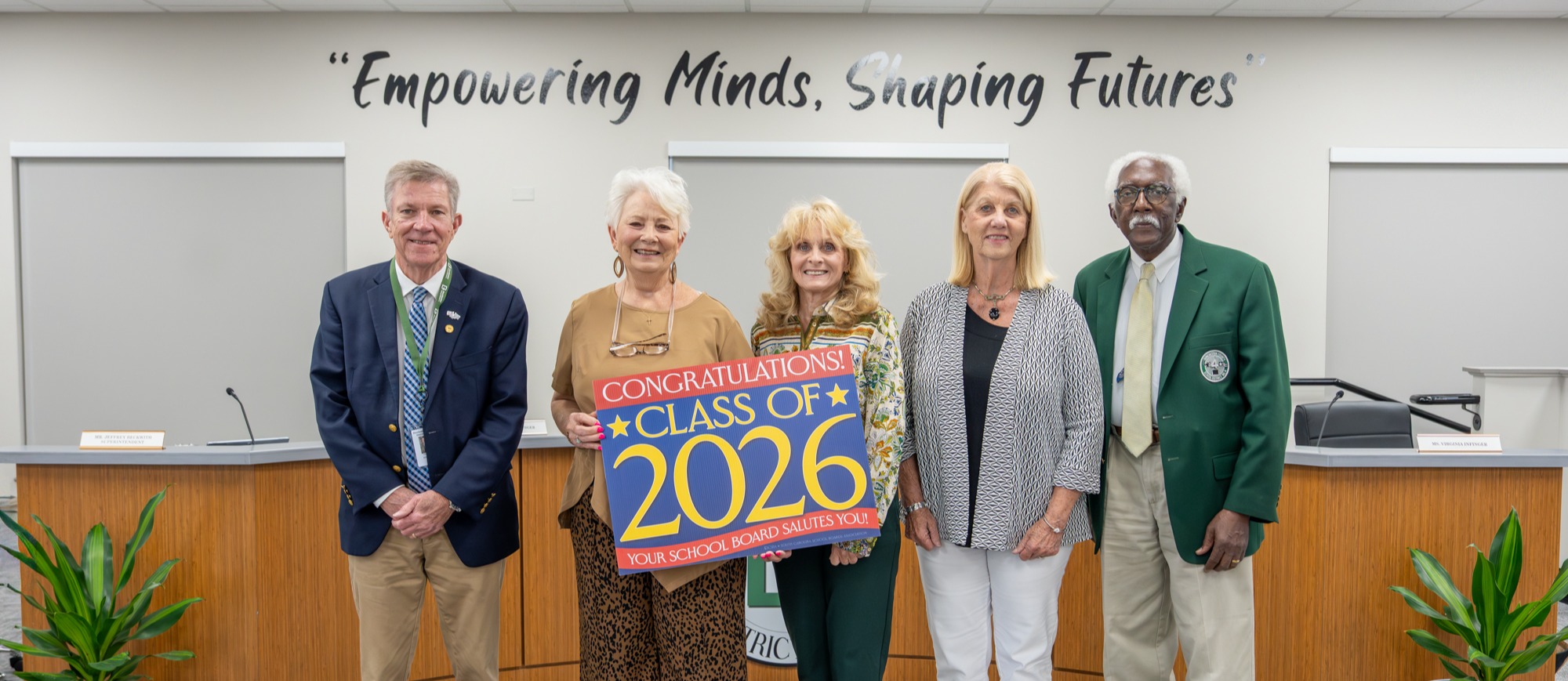 Five members of the Dorchester School District Four Board of Trustees stand in the boardroom under the district motto, “Empowering Minds, Shaping Futures.” Two board members hold a sign congratulating the Class of 2026 that reads, “Your School Board Salutes You!”