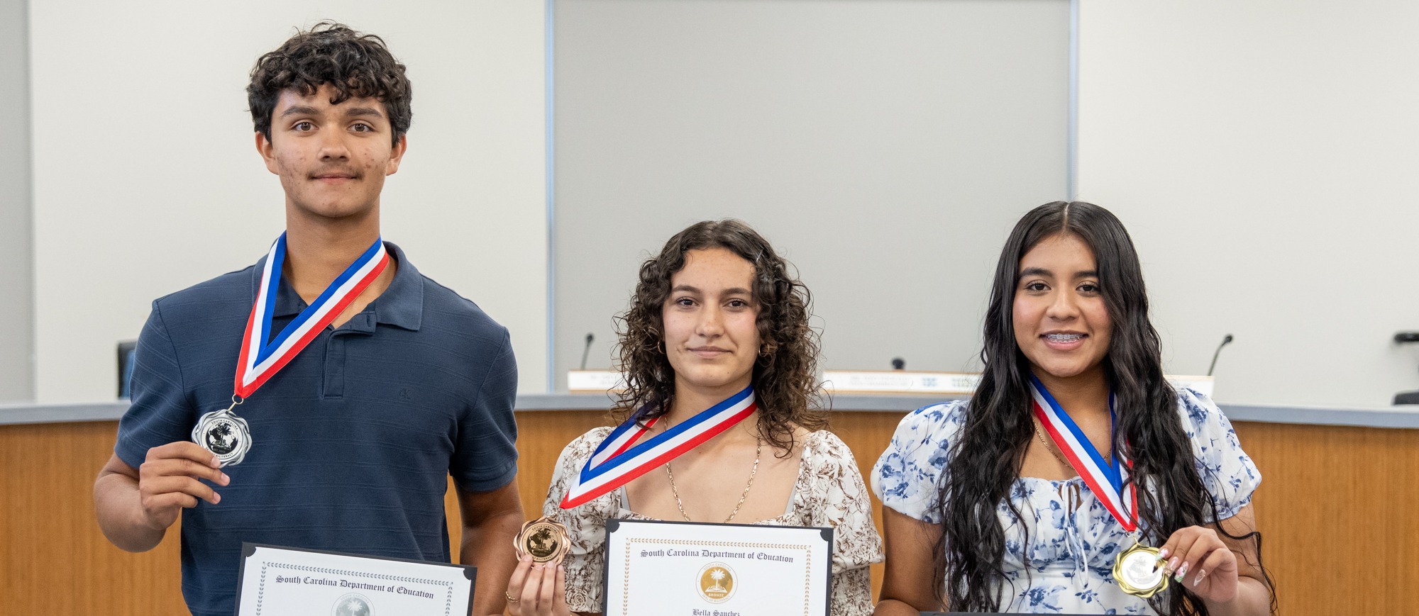 Three students stand in a school board room holding South Carolina Seal of Biliteracy certificates and medals. The student on the left wears a navy polo and gray pants, while the two students on the right wear floral dresses. A wall behind them displays the phrase “Empowering Minds, Shaping Futures.”