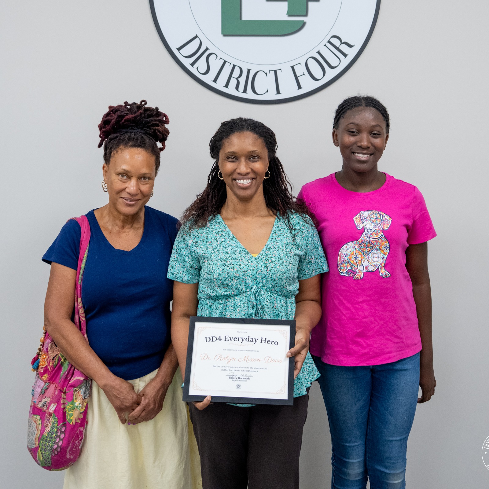 Dr. Robyn Mixon-Davis stands with two family members beneath the Dorchester School District Four logo while holding a DD4 Everyday Hero certificate. One woman stands beside her carrying a colorful shoulder bag, and a young girl in a bright pink shirt stands on the other side smiling for the photo.